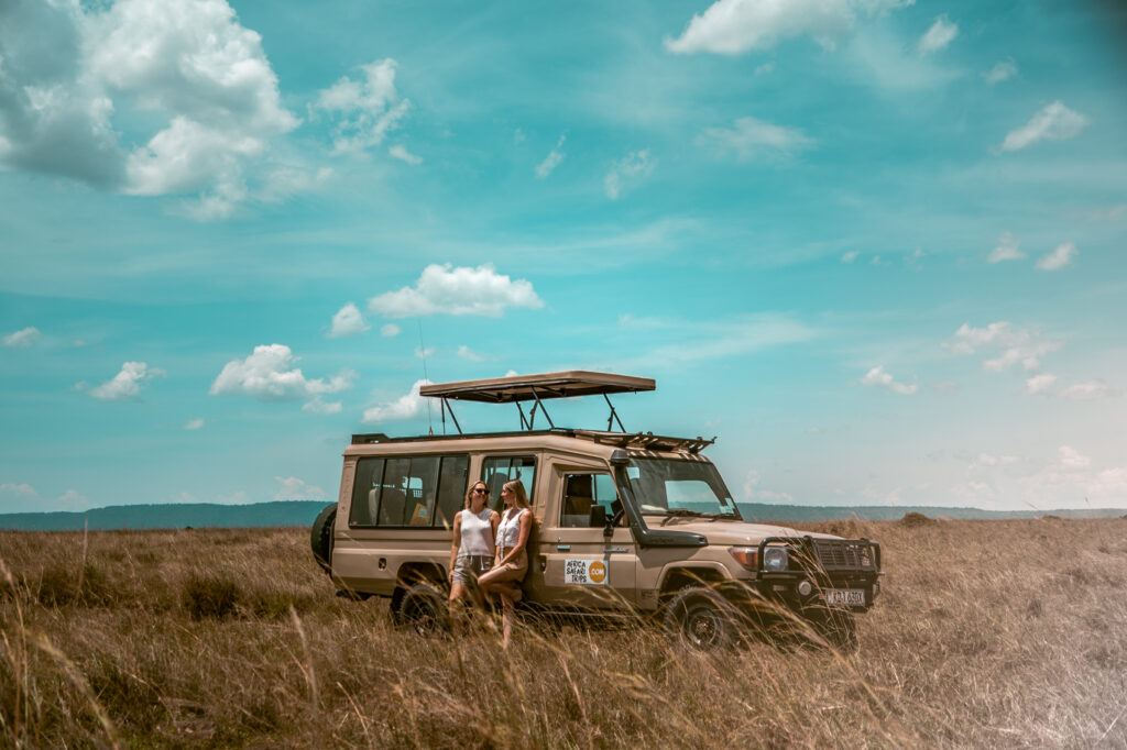 To kvindelige rejsende hyggesnakker foran en safari-jeep på en ellers øde savanne.