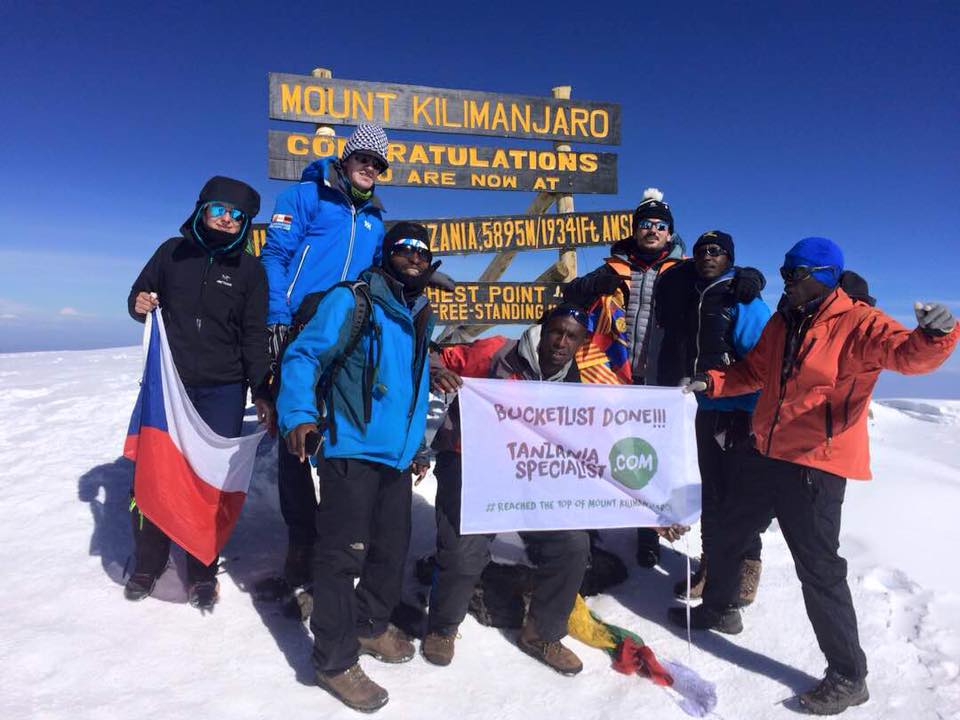 Group of people in winter clothes standing on the summit of Kilimanjaro in front of a sign with a flag of Tanzania Specialist